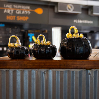Three black glass pumpkins with iridescent yellow stems on a wooden surface in a hot shop setting.